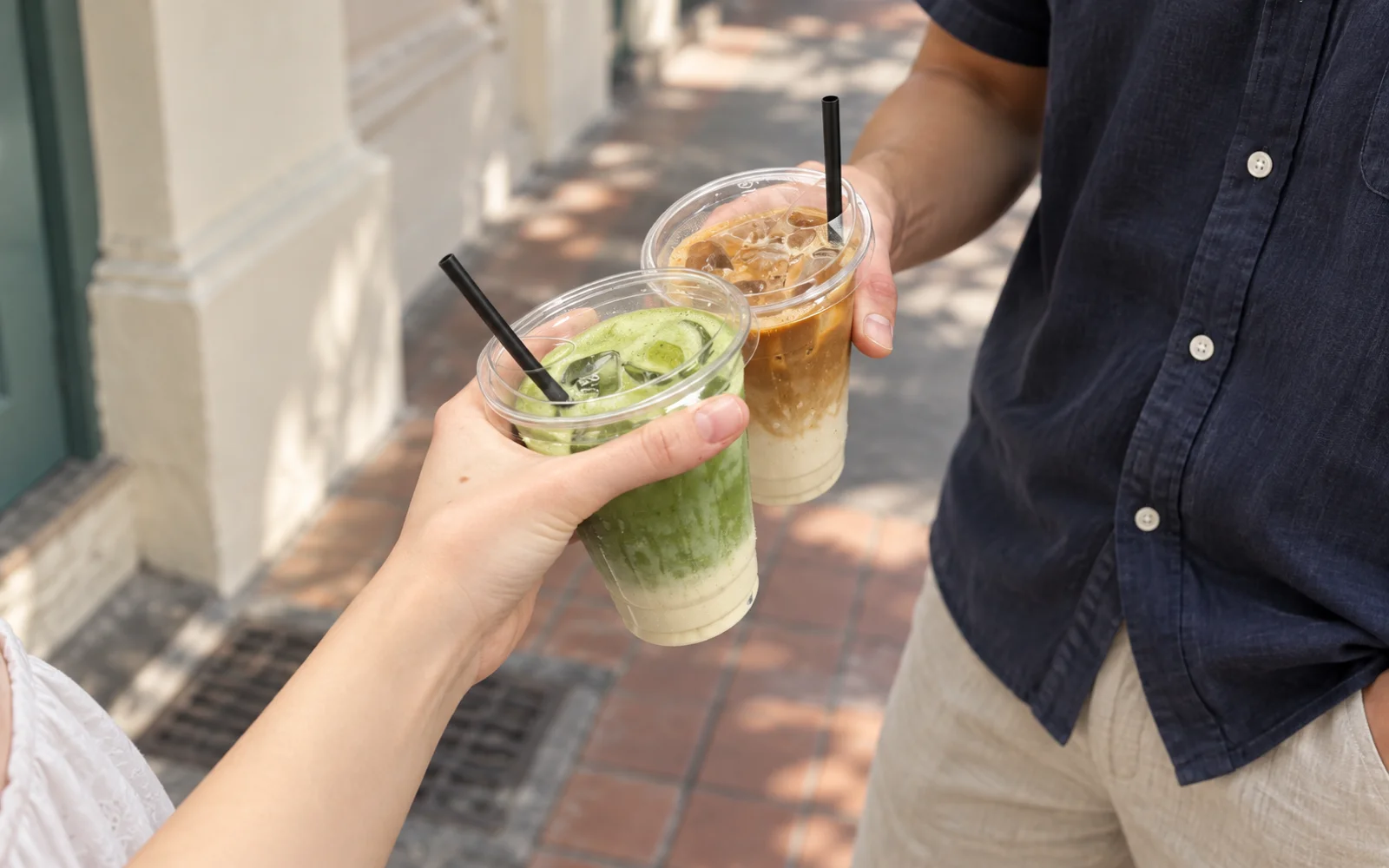 Two people holding iced matcha and coffee on a sunlit walkway